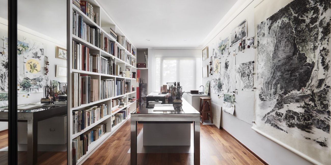 A well-lit room with wooden flooring, shelves filled with books, a desk with art supplies, and large paintings on the walls.