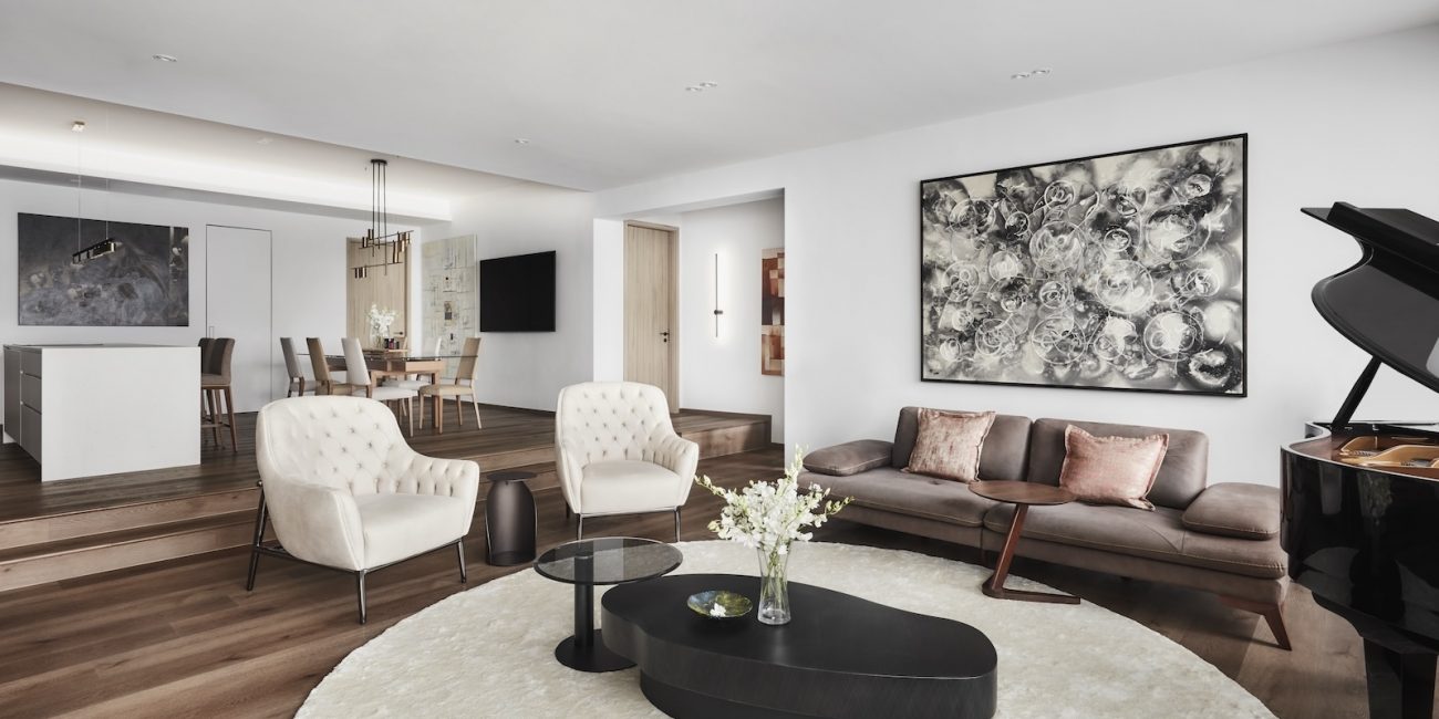 Modern living room featuring a grand piano, a round rug, a brown sofa with cushions, two beige chairs, and abstract artwork. A dining area and kitchen are visible in the background.